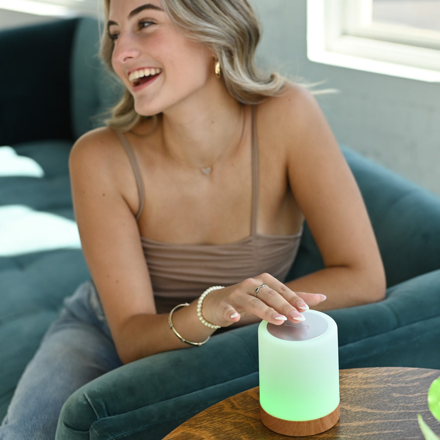 Woman smiling while touching a glowing green friendship lamp on a wooden table in a modern living room.